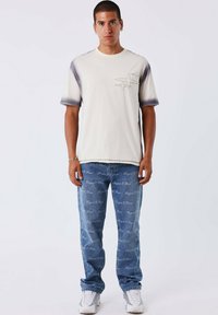 T-shirt color crema con dettagli blu scuro, dotata di una tasca decorativa. Abbinata a jeans di denim blu chiaro, con stampa di testo bianco.
