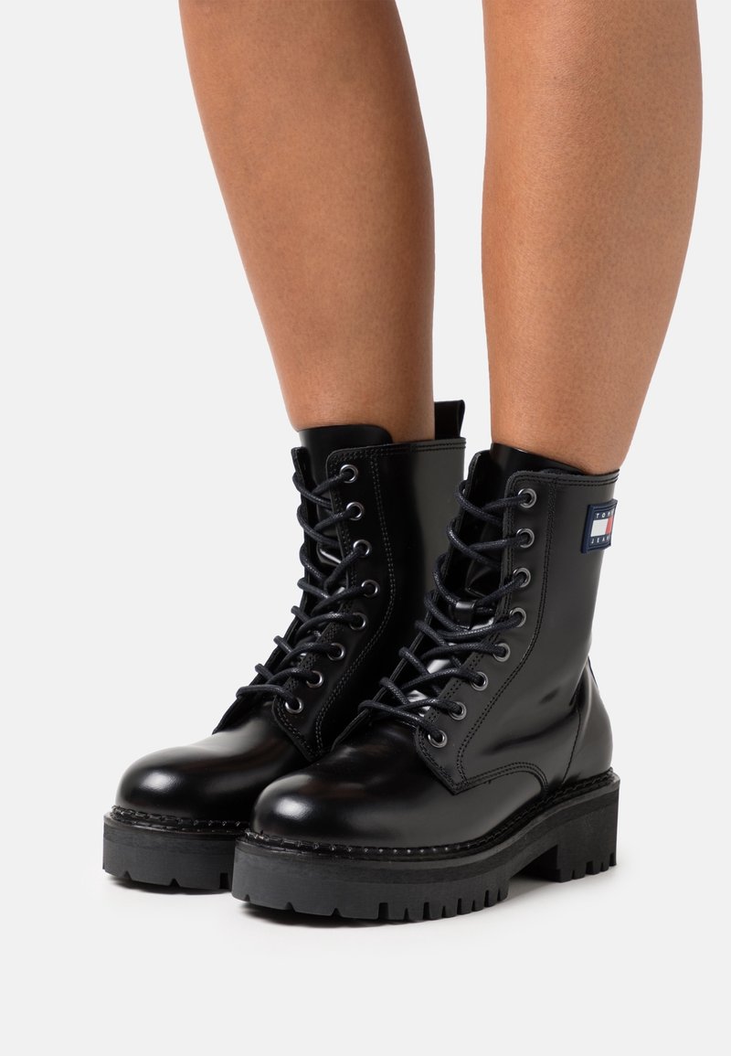 Bottes de combat noires à lacets avec semelles épaisses portées sur des jambes nues, comportant un petit patch logo rectangulaire sur le côté supérieur extérieur.