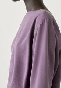 Haut à manches longues en tissu lavande doux et texturé. Comprend un col rond et une coupe décontractée, mettant en valeur un drapé aux épaules.