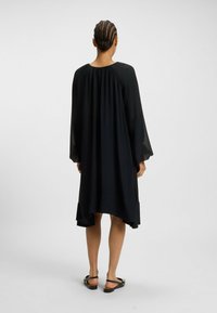 Svart flödande klänning med långa genomskinliga ärmar, rund halsringning och hi-low fåll. Slät textur och lättviktsmaterial. Enkelt design.