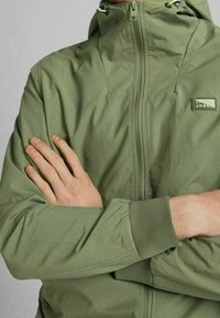 Giacca zip-up verde chiaro con colletto alto, polsini elasticizzati e un logo discreto sul petto. Tessuto liscio con finitura opaca.
