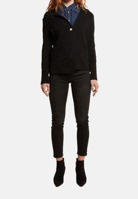 Czarny kardigan z okrągłym kołnierzem i zapięciem na pojedynczy guzik, noszony na niebieskiej dżinsowej koszuli, zestawiony z czarnymi obcisłymi dżinsami i botkami za kostkę.