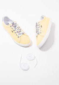 Canvas sneakers in pale yellow with white soles, featuring decorative laces and logo accents. Includes extra white laces.
