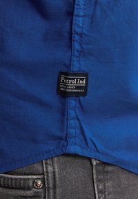 Modrá látka s hladkým povrchem, s částečně viditelnou etiketou "Petrol Ind" vedle šedého denimu. Detaily šití na lemu.