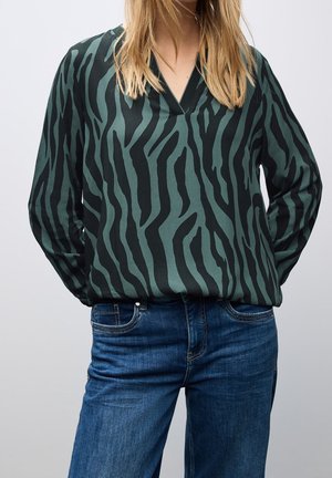 Frau trägt eine schwarz-dunkelgrün gestreifte Bluse mit langen Ärmeln im Zebra-Muster, die in blaue Jeans aus Denim gesteckt ist, mit den Händen hinter dem Rücken.