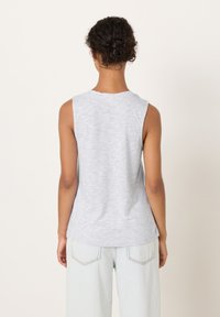 Žena s kovrčavom kosom skupljenom u punđu, odjevena u svijetlosivu majicu bez rukava i svijetloplave traperice, stoji okrenuta leđima prema jednostavnoj pozadini.