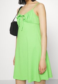 Grön klänning i ett texturerat, lätt tyg, med figurnära liv och en rosett framtill, ihop med en liten svart texturerad väska.