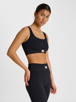 Kvinde med mørkt hår iført en sort ribbet sports-bh og matchende højtaljede leggings, stående mod en ensfarvet lys baggrund.