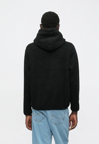 Άνδρας που φορά μαύρη φλις μπλούζα με κουκούλα και ανοιχτό μπλε τζιν, στέκεται με την πλάτη στραμμένη προς την κάμερα, μπροστά σε λευκό φόντο.