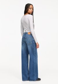 Breda blå jeans i en medeltvätt, med hög midja, två bakfickor och en beige läderetikett ovanför den högra fickan. Långärmad grå topp.