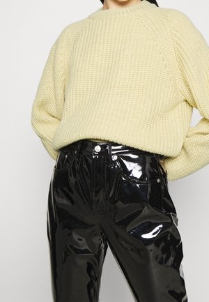 Personne portant un gros pull en maille crème rentré dans un pantalon en cuir verni noir brillant taille haute, avec poches avant et passants pour ceinture.