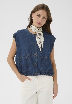 Mlada ženska, oblečena v brezrokavnik iz modrega jeansa z gumbi, bele hlače, biserne uhane in belo rutico z belimi pikami na vratu, pozira z eno roko v žepu.