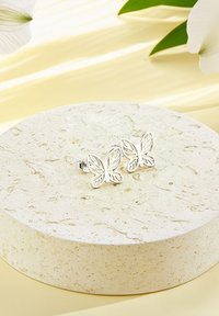 Silverfärgade fjärilsörhängen med intrikata utskärningsmönster, visade på en cirkulär texturerad stonyta mot en mjuk gul bakgrund.