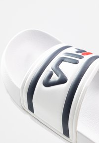 Vit halkskyddssandal med en strukturerad rem, som har en framträdande svart logotyp, röd accent och marinblå ränder längs sidan.