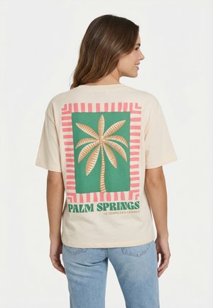 Donna che indossa una t-shirt beige con una grande grafica di una palma e la scritta "Palm Springs" sul retro, abbinata a jeans azzurri.