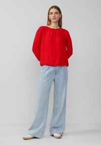 Un blouse rouge à manches longues avec un col rond et des détails froncés aux épaules, assorti à un jean à jambes larges bleu clair et des chaussures beiges.