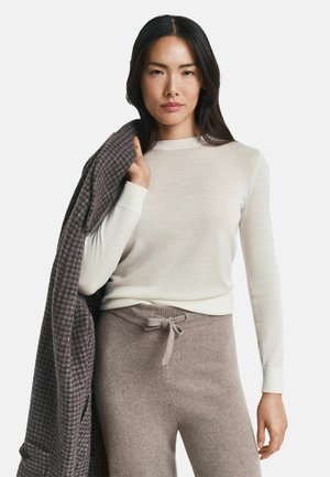 Vrouw in een crèmekleurige lange mouwtop en taupe gebreide broek, met een opgevouwen grijs houndstooth jas over haar schouder tegen een witte achtergrond.