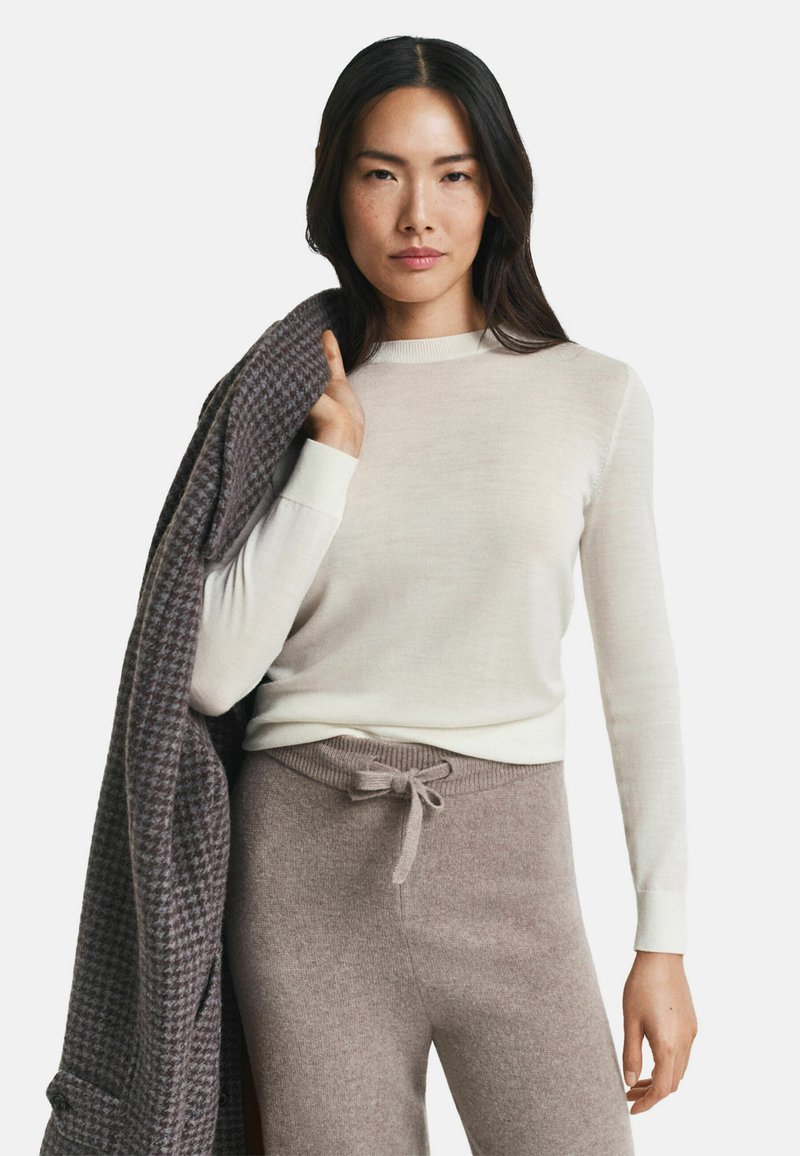 Donna che indossa un top a maniche lunghe color crema e pantaloni a maglia color taupe, tiene un cappotto piegato grigio a scacchi sulla spalla su uno sfondo bianco.