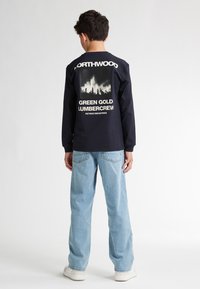 Camicia a maniche lunghe blu navy con stampa grafica bianca sul retro, che presenta montagne e alberi. Abbinata a jeans azzurri chiari, dal taglio morbido.