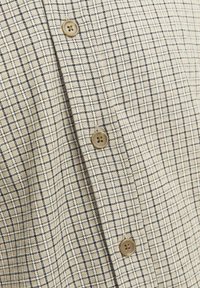 Chemise beige avec un motif en grille de lignes navy, ornée de boutons marron et d'un tissu texturé. Accent sur le design structuré et le détail des boutons.