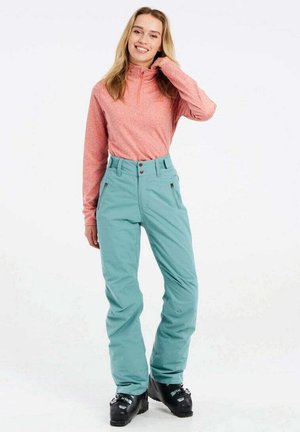 Light pink long-sleeve pullover and teal ski pants with a high waist and two side pockets, paired with black ski boots.