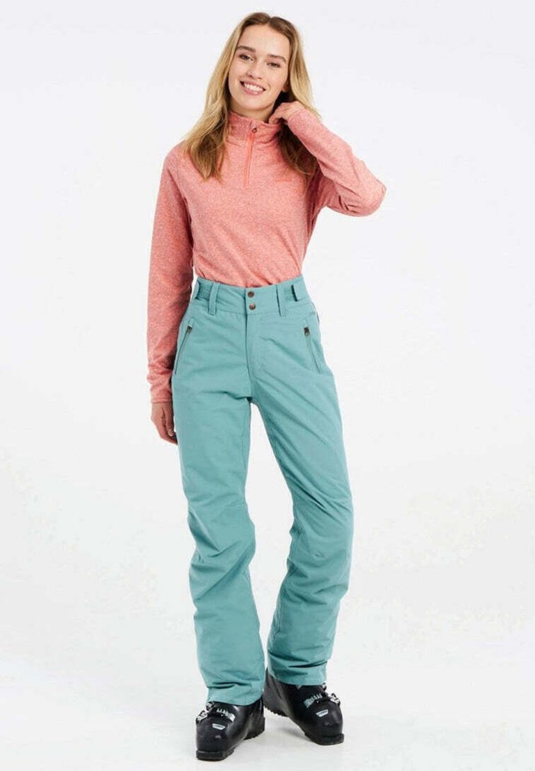 Lichtroze lange mouwen pullover en teal skibroek met een hoge taille en twee zijzakken, gecombineerd met zwarte skischoenen.