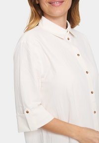 Camicia bianca con colletto, maniche arrotolate e bottoni marroni, realizzata in un tessuto testurizzato.