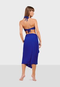 Blå halterneck bikiniöverdel med knytning bak och matchande inbärande kjol. Slitstark, lättviktsmaterial med flödande textur och asymmetrisk nederkant.