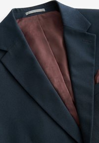 Marineblazer van een textuurstof, met een bordeauxrood patroon in de voering en een notch lapel. Pochette en interieurdetails zijn zichtbaar.