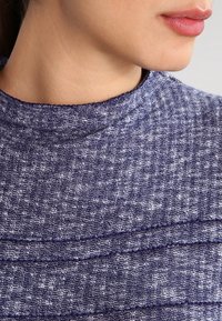 Texturerad marinblå tyg med hounds-tooth mönster, med hög halsringning och horisontella sömdekorationer. Mjukt, vävt material.