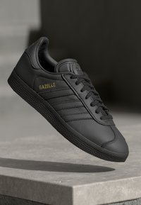 Juodos odinės sportinės bateliai su tekstūruota juoda gumine baze, turintys tris šonines juosteles ir "GAZELLE" auksine spalva viršuje. Klasikinis žemas dizainas.