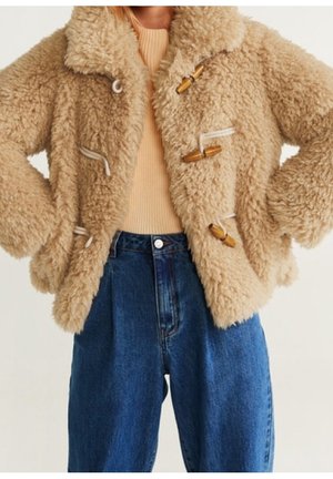 Kobieta nosząca beżowy sztuczny kożuch typu toggle na prążkowanym brązowym topie i niebieskich dżinsach z wysokim stanem.