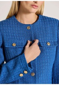 Blauwe geweven jas met textuurstof, gouden knopen en opgestikte zakken. Ronde halslijn met een gestructureerde pasvorm en gedetailleerde manchetten.