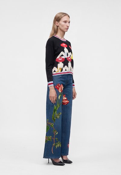 Femme portant un pull noir avec un motif de chapeau rouge et un jean bleu large avec une grande broderie florale, debout de profil sur fond blanc.