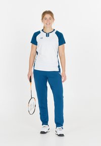 Vitt och marinblått träningsoutfit med korta ärmar, blåa byxor och ett mönstrat badmintonracket. Materialet verkar lätt och sportigt.