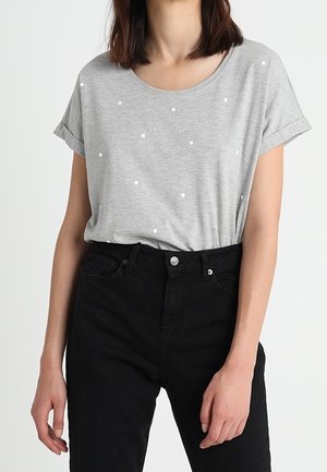 Kobieta ubrana w jasnoszarą koszulkę z krótkim rękawem w drobne białe kropki oraz wysokiej talii czarne spodnie na jednolitym tle.