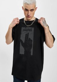 Sort oversized t-shirt med et grafisk print af en hånd i grå nuancer, placeret på øverste brystområde, med en kædetrøje og synlig tattoo på modellens hånd.