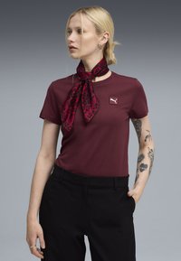 Mørkerød bomulds T-shirt med rund hals og broderet logo. Parret med sorte bukser og en mønstret rød og sort tørklæde bundet om halsen.