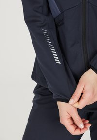 Marinblå softshell-jacka med dragkedja fram, med vita reflexdetaljer och en åtsittande mudd. Slät textur med en något glänsande yta.