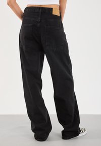 Černé džíny s volnějším střihem, se dvěma zadními kapsami, hnědým koženým štítkem na pase a hladkou texturou.