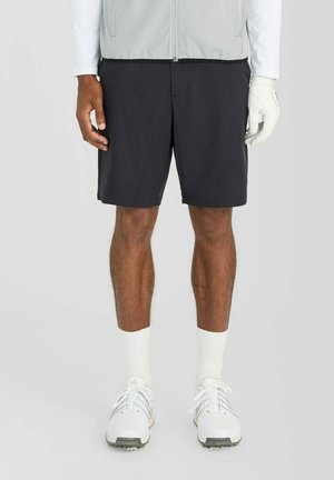 Mann trägt schwarze Shorts, weißen Golfhandschuh an der rechten Hand, weiße Socken und weiße Sportschuhe und steht vor einem einfarbigen Hintergrund.