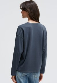 Langärmliges Hemd aus dunkelgrauem Stoff. Mit lässigem Schnitt, Rundhalsausschnitt und sichtbaren Nähdetails. Kombiniert mit hellblauen Jeans.