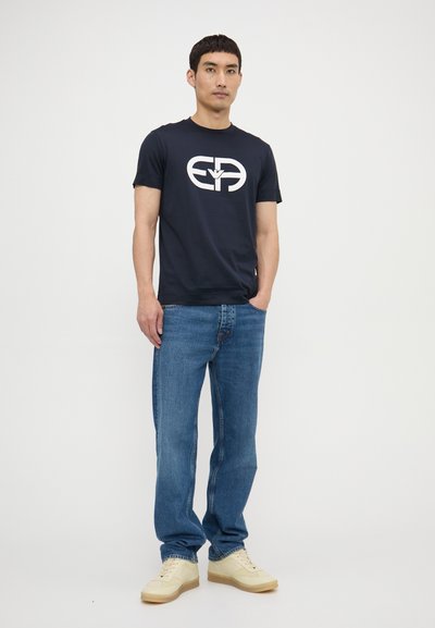 Tricou din bumbac de culoare bleumarin cu un design alb, asortat cu blugi decolorati de culoare albastră și tenisi galbeni, stând în fața unui fundal simplu.
