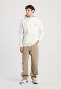 Felpa bianca con cappuccio e tasca a marsupio, decorata con il logo "Rocky Mountains". Abbinata a pantaloni beige a vestibilità ampia e sneaker grigie.