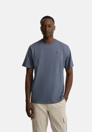 Hombre que lleva una camiseta azul lisa de manga corta y pantalones cargo beige, de pie con una mano en el bolsillo sobre un fondo blanco.