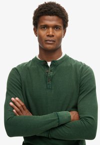 Mężczyzna o kręconych włosach i poważnym wyrazie twarzy, ubrany w zieloną bluzę Henley z długim rękawem, ze złożonymi ramionami, stojący na tle jednolitej białej ściany.