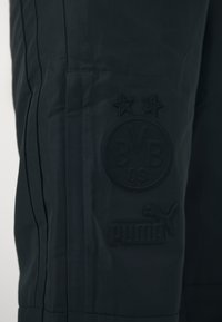 Čierne športové nohavice s textúrovanou látkou, s vystúpeným čiernym logom "BVB 09" a značkou Puma na dolnej časti.