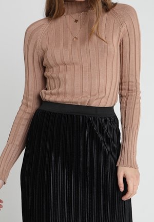 Žena oblečená v béžovém žebrovaném svetru s dlouhými rukávy a černé plisované sametové sukni s vysokým pasem, zdobené vrstvami zlatých náhrdelníků.