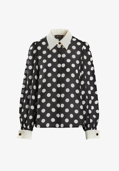 Melna blūze ar baltām punktiņām. Iezīmējas ar klasisku apkakli, uzpūstām piedurknēm un baltām aprocēm ar apaļām zelta akcentiem. Gluda tekstūra.