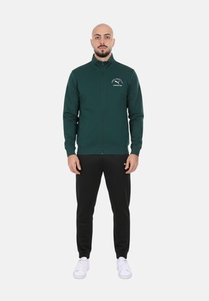 Uomo in piedi, volto frontale, indossa una giacca con cerniera verde scuro, pantaloni neri e scarpe da ginnastica bianche su uno sfondo chiaro semplice.